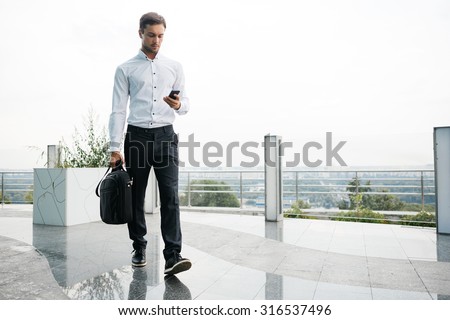 Young businessman is walking with his case. He is writing a message on his mobile phone. Skyline on a background