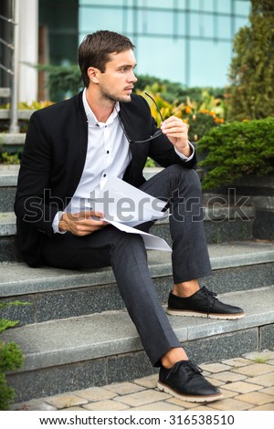 Elegant man is keep watching over something he interested. He is sitting on the stairs in dress code. He is keeping his notes on his hands.