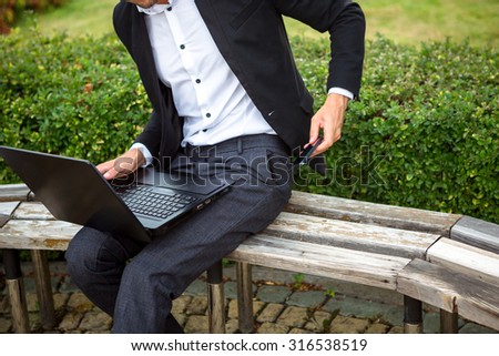 Fashionable man in the street is sitting on the bench with note book and phone. He wears a nice dress code. He is working.  Green bushes around him.