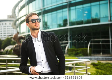 Man in dress code is standing near the handrail. He is drinking his coffee. He is wearing a black glasses. He is looking for somewhere.