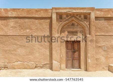 A traditional arched doorway with gypsum carvings and a carved door stands in bright sunlight in a wall of Riffa Fort, Bahrain, in the Arabian Gulf.