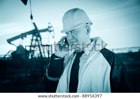 Oil worker in uniform and helmet, with mobile phone on of background the pump jack and sky. Toned.