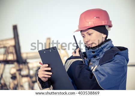 Female worker in the oil field talking on the radio wearing red helmet and blue work clothes. Industrial site background. Toned.
