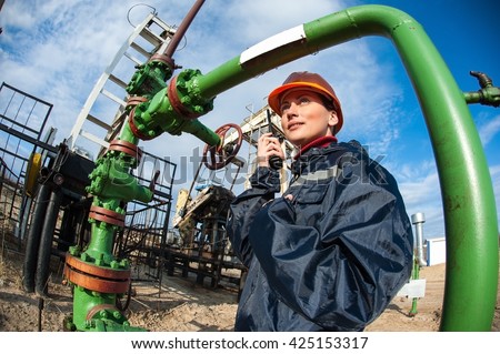 Female worker in the oil field talking on the radio wearing orange helmet and blue work clothes. Industrial site background.