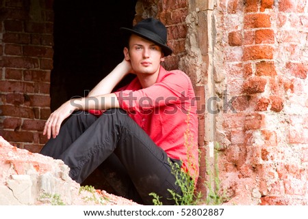 Elegant fashion man model wear red shirt and hat near bare walls. Handsome young man look. Young casual man portrait wear a hat