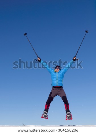 The man in snowshoes and with trekking poles jumps in the mountains on the background of blue sky.