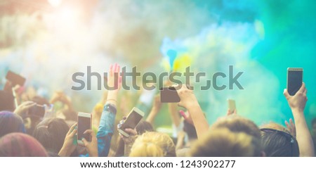 Crowd of people dancing and celebrating Holi festival of colors. People taking photos with mobile phones at color festival