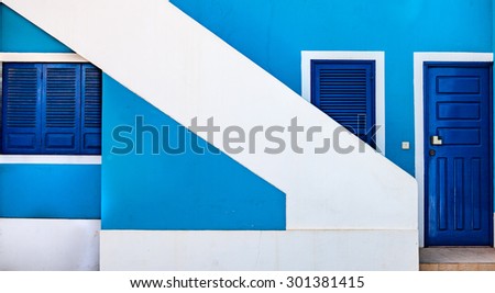 A house front with a vivid blue facade, two marine blue doors and one window with marine blue, wooden shutter. White stairway going diagonally through the image.