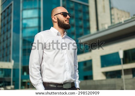 Businessman with beard in sunglasses in white shirt standing near office building