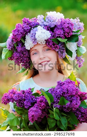 Portrait of a little girl with a bouquet of flowers in her hands and a wreath of flowers of lilac color . The girl is smiling and looking at the camera.  The photo was taken outdoors.