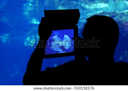 Photographing a jellyfish aquarium.\
Silhouette of a man photographing a jellyfish aquarium with a tablet