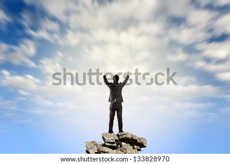 Successful businessman stand on stone and open arms against dramatic cloudy sky.