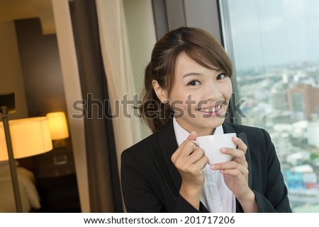 Asian business woman holding a cup of coffee and looking into distance near the window in hotel room.