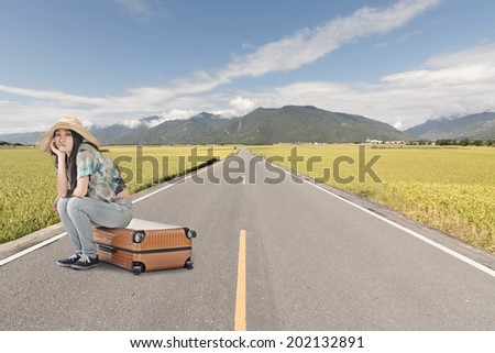 Asian woman thinking and sitting on a luggage on the ground of road in countryside.