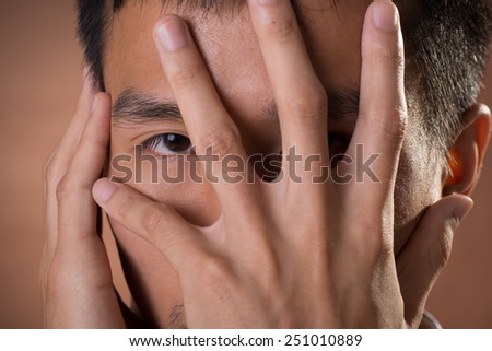 Young Chinese frank businessman cover his face by hand and peeking through finger, close up portrait in studio.