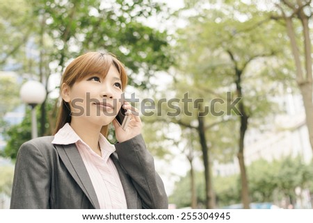 Attractive Asian business woman talk on phone under green tree in modern city, closeup portrait shot at Xinyi business district, Taipei, Taiwan.