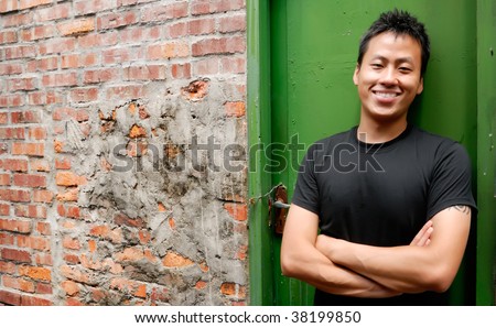 There is an Asian man stand against a old door and smile.