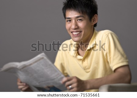Happy smiling young man of Asian sit and reading newspaper over gray studio background.