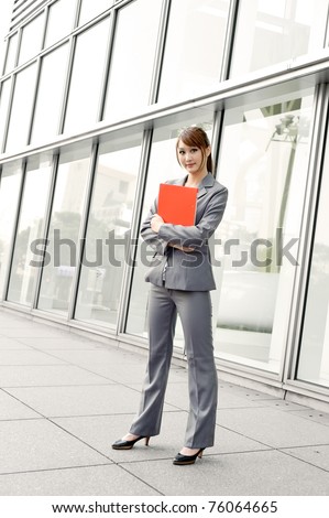 Young smiling business woman holding red file document and standing looking at you, full length portrait outside of modern buildings.