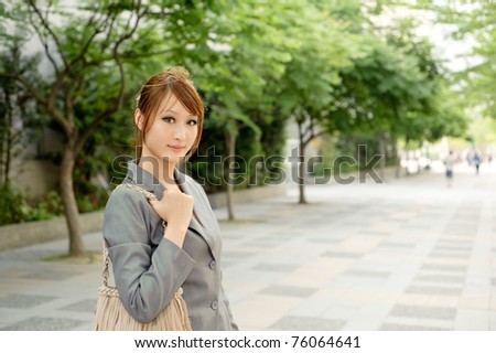 Young business woman walking on city street with green trees and looking at you, half length closeup portrait outside of modern buildings.