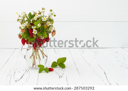 Bouquet of wild strawberries in a glass jug on white old wooden background