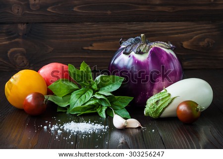 Purple and white eggplant (aubergine) with basil, garlic and tomatoes on dark wooden table. Fresh raw farm vegetables - harvest from the garden in rustic kitchen. Rural still life