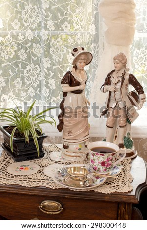 Vintage porcelain figures , bone china tea cup and sterling silver tea strainer on vintage magazine stand. Lace embroidery table cloth.