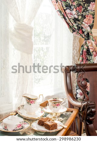 Afternoon tea. Vintage style interior design. Matching patterns of tableware and the curtain. Vintage fine bone china, silver tea strainer, antique rocker chair. Cross stitch tea tray.