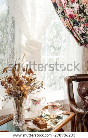 Afternoon tea. Vintage style interior design. Matching patterns of tableware and the curtain. Vintage fine bone china, dried flowers, silver tea strainer, antique rocker chair. Cross stitch tea tray.