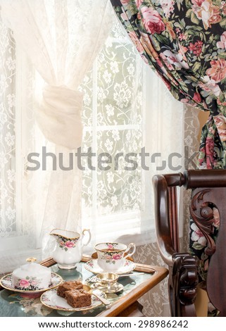 Afternoon tea. Vintage style interior design. Matching patterns of tableware and the curtain. Vintage fine bone china, dried flowers, silver tea strainer, antique rocker chair. Cross stitch tea tray.