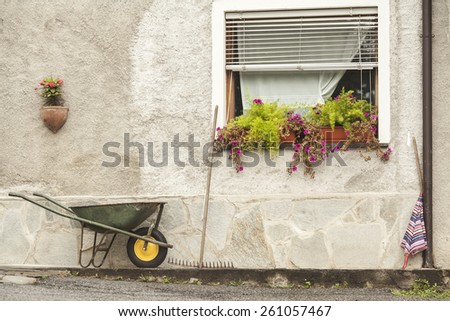 rural scene of creative house wall showing open window with flowers in pots, umbrella, garden trolley and rake