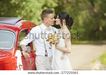 Happy newly wed couple looking at each other with love next to a red vintage car in park. bride holding a bouquet and groom hugging his wife