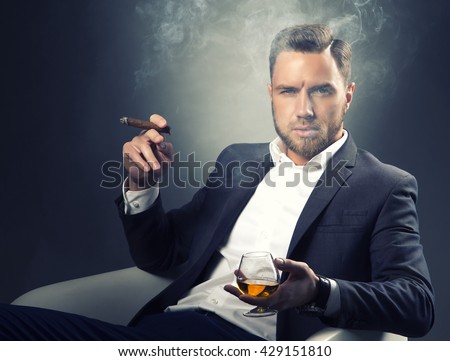 Young handsome bearded caucasian man sitting on chair with cognac and a cigar. Perfect skin and hairstyle. Wearing grey suit and watch. Studio portrait on gradient black to grey background. Toned