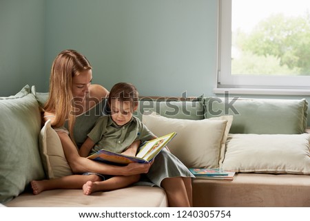 Young mother, read a book to her child, boy in the living room of their home, rays of sun going through the window