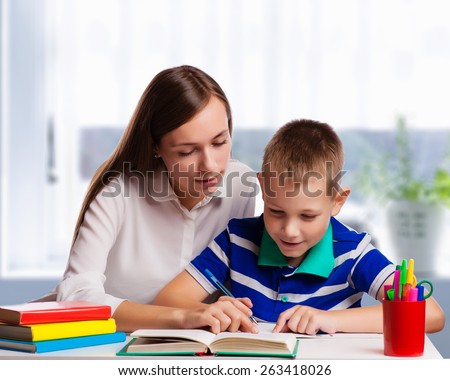 Young mother sitting at a table at home helping her small son with his homework from school as he writes notes in a notebook