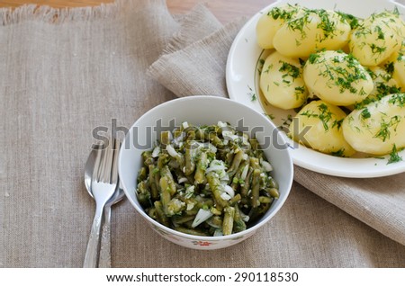 Marinated green beans with onions, garlic, dill. Cooked with olive oil. In the background a bowl with boiled potatoes