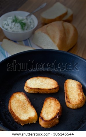 Toasts in a pan. Bowl with cottage cheese and greens, sliced bread in the background.