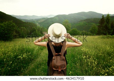woman traveler with backpack holding hat and looking at amazing mountains and forest, wanderlust travel concept, space for text, atmosperic epic moment