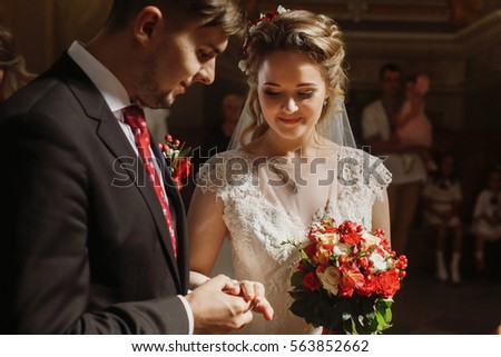 Romantic couple exchanging rings during wedding ceremony in church, handsome groom putting on wedding ring on beautiful bride\'s hand closeup