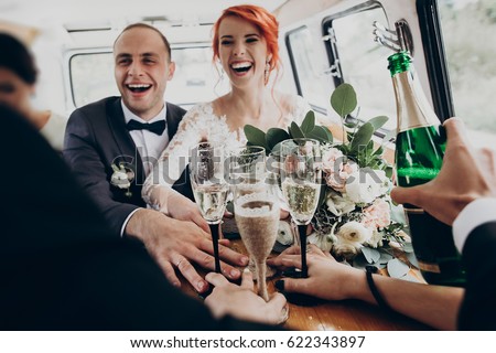 stylish happy bride and groom toasting with glasses of champagne and having fun with bridesmaids and groomsmen inside of retro car. emotional moment, space for text. wedding party