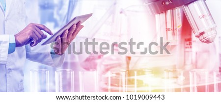 Science lab research and development concept - Scientist holding tablet computer with scientific instrument, microscope and chemical test tube in laboratory background.