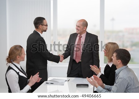 Two businessmen shake hands. A group of people formed of women and men at the table applauds them.