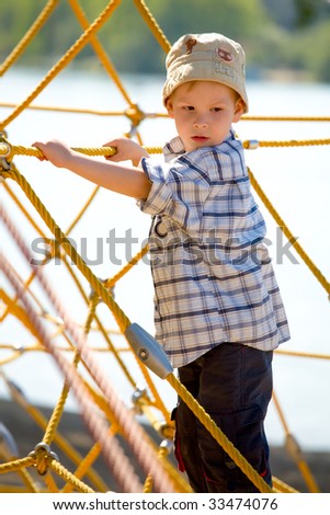 Boy on playground next to lake, climbing net of yellow ropes.