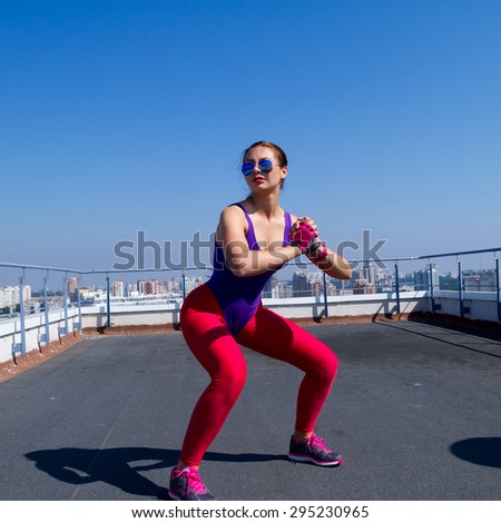 Fitness girl exerting outdoors going jump squat in amazing top city view. Fit female woman athlete cross-training outside. Wearing wearing sportive violet swimsuit,pink leggins and sunglasses.