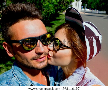 Happy young couple in love kissing and taking self-portrait while the walking in the city park. Trendy romantic couple in similar outfit wearing jeans vest, swag cap hat and mirrored sunglasses.