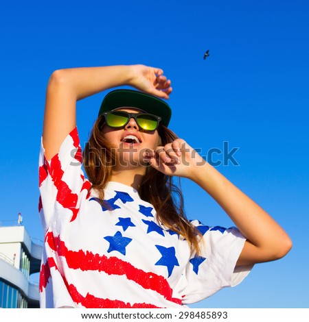 Summer fashion portrait of young teen girl having fun go crazy, wearing trendy top with american flag print,hat, hipster mirrored glasses. Sitting on the terrace with amazing sea view. Joy Sea Sunset