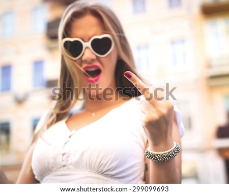 Fashion swag sexy blonde young woman showing middle finger one hand Wearing white dress,pink lips, heart shaped  sunglasses with city street background.Young sexy girl showing middle finger gesture.