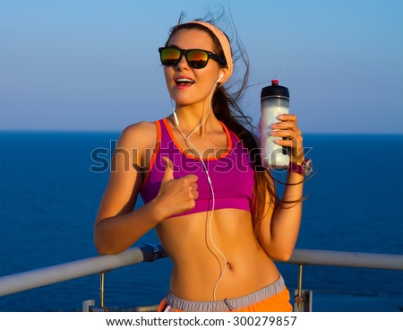 Fitness beautiful woman drinking water and sweating after exercising on summer hot day near the sea. Female athlete after work out. Wearing bright purple sport bra,orange sport shorts,mirrored glasses