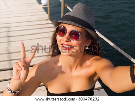 Young smiling woman in hat and sunglasses making selfie with phone. Happy woman taking selfie photo with smartphone camera summer travel vacation to the coast. wearing black top corset grey hat