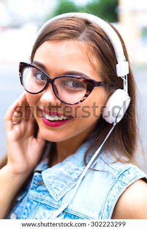 Close up fashion portrait of young pretty woman listening her favorite music in white headphones. Stylish teenage hipster girl in earphones, wearing jeans vest,brown clear glasses.Smiling teenage girl